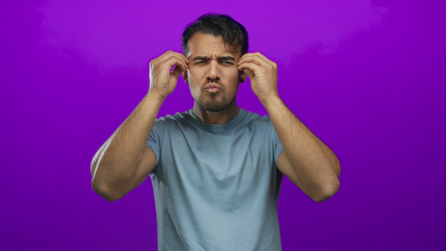 Young hispanic man appears confused against an isolated purple background, expressing stress through raised hands and an uncertain gaze, wearing a light blue t-shirt.