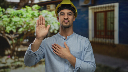 Naklejka premium Young man in hardhat making oath gesture outdoors in green park setting with trees in background.