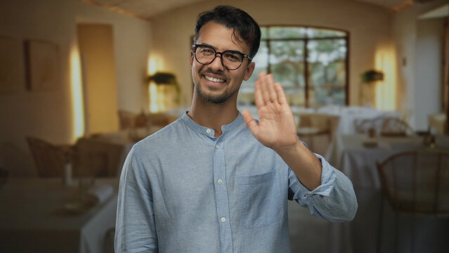 Young man with glasses smiling and waving hello in a cozy indoor restaurant setting featuring coffee and relaxed ambiance, highlighting a warm and friendly atmosphere.