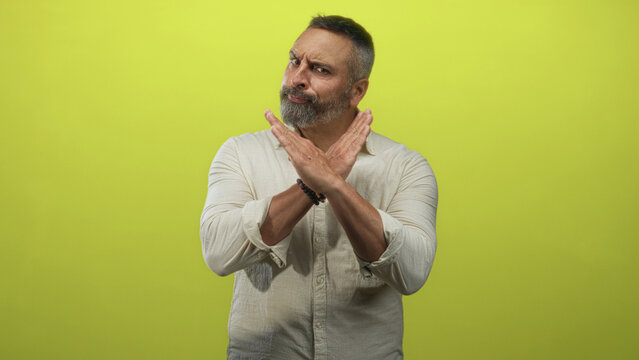 Middle aged hispanic man with grey hair and beard crossing forearms in an x gesture in lime green studio; rejection caution boundary.