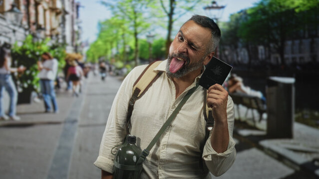 Man middle aged winking and sticking out tongue holding passport on city street while wearing backpack and canteen; playful wanderlust.