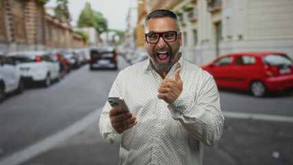 Man taps smartphone with fingers on a busy city street wearing glasses and beard, smiling at...