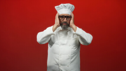Middle aged man chef in white uniform touching head with hands, wearing hat and glasses in red studio; kitchen stress concern.