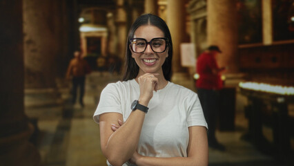 Woman wearing oversized glasses with hand on chin and folded arm smiling in a church building lit by candles and columns; reflective serene.