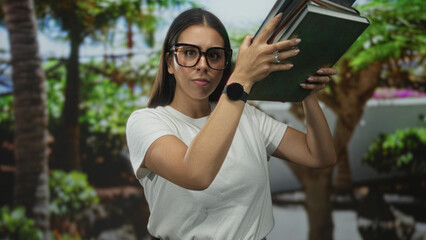 Woman wearing large glasses and white t shirt holding a stack of hardcover books in park among trees and palms; study concentration.