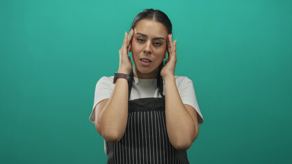 Young brunette woman in striped apron touching temples with closed eyes and slight grimace in teal studio; stress headache tension.