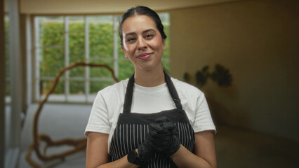 Woman in striped apron and black gloves smiling with gloved hand on neck in studio windowed room with sculpture and plant; serenity hospitality.