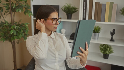 Woman holds tablet and touches hair while seated by bookcase and potted plants in an office building; focus productivity.