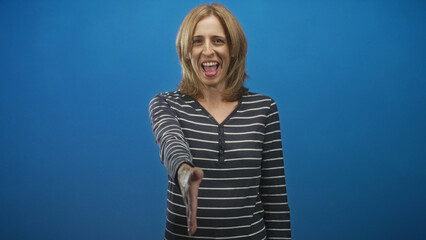 Woman smiling extends right hand for handshake in studio blue backdrop wearing striped top and natural smile; friendly greeting.