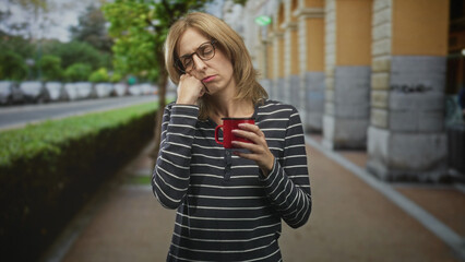 Fototapeta premium Woman holding red mug with hand on cheek, wearing glasses and striped shirt, standing on street sidewalk near building columns; tired solitude.