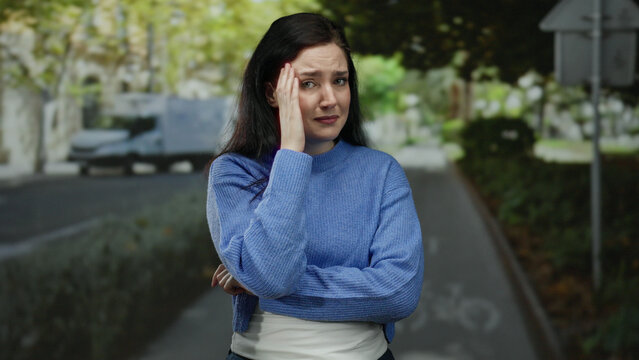 Woman standing on a bustling street wearing a blue sweater outdoors looking distressed and emotional with blurred vehicles in the background creating a tense urban atmosphere.