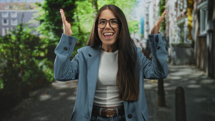 Young woman wearing glasses and a blue blazer raises both hands with an open mouth in an expressive gesture on a street lined with trees and bollards; surprise tension.