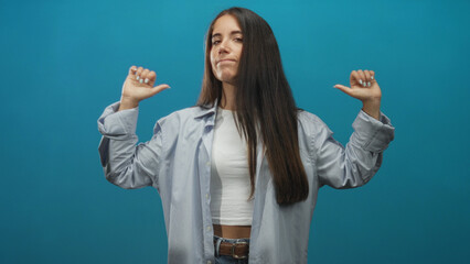 Woman with long straight hair, thumbs pointing to self, wearing white crop top, open shirt and jeans against blue studio backdrop; confidence.