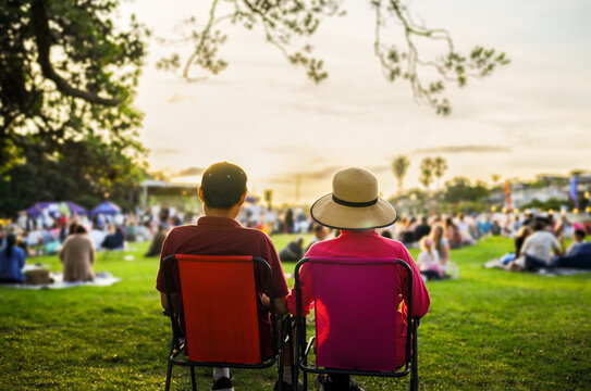 Couple sitting on the picnic chairs, listening to the music concert. Unrecognizable crowd of people in the park.