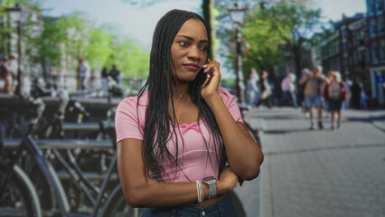 Woman holds phone to ear on a street, arm crossed and listening beside parked bicycles and buildings, wearing pink top and bracelets; thoughtful reflection.
