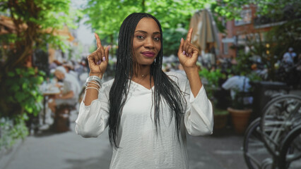 Woman pointing both index fingers upward at a busy street cafe, visible fingernails, braided hair and white blouse; confidence empowerment.