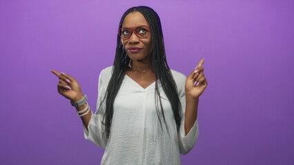 African woman pointing index fingers upward with long braided hair, red glasses, lilac nails and bracelets in studio; playful confidence.