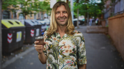Fototapeta premium Man with long blond hair smiles while holding a traditional mate cup on a cobblestone street in sunlight; serenity.