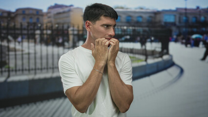 Man in white shirt biting fingernails with tense hands in front of urban building fence on busy street; anxiety.