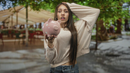Young woman holding pink piggy bank with bare right hand, offering it forward while touching hair and looking concerned in street market; saving anxiety.