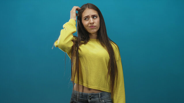 Teenage girl scratching head with hand, wearing yellow sweater and jeans in studio with teal backdrop; confusion uncertainty.