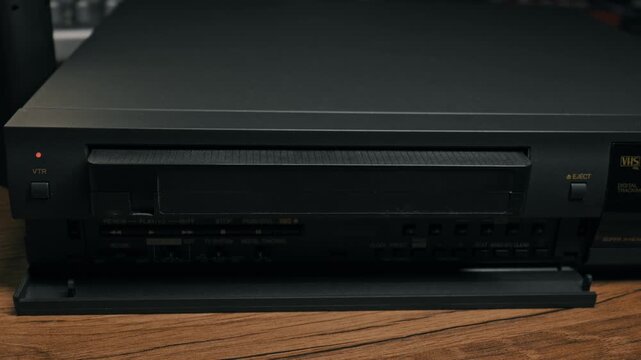 Male hand presses eject button on black vintage VCR player to release cassette. Person removes analog tape from retro video recorder on wooden surface.