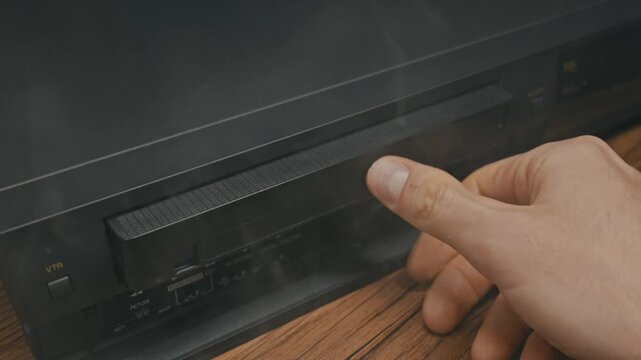 Male hand inserts black VHS cassette into player then ejects it after pressing button. Atmospheric smoke swirls around vintage electronic equipment on wooden surface.
