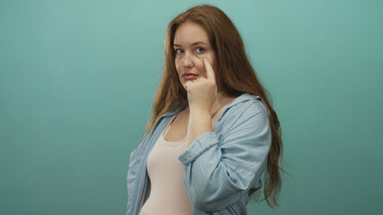 Young caucasian woman pointing finger to eye with direct gaze in teal studio; alertness observation.