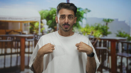 Fototapeta premium Man in white t shirt points hands to chest and raises eyebrows on a sunny terrace by wooden railing; surprise.