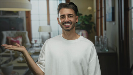 Obraz premium Man wearing white shirt shows open palm and smiles broadly beside sofa in modern building; hospitality warmth.