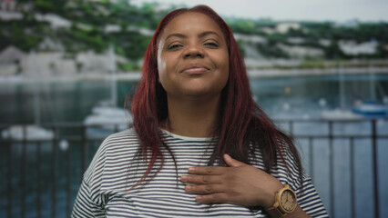 Woman with long red hair touches chest and smiles beside seaside building overlooking boats on...