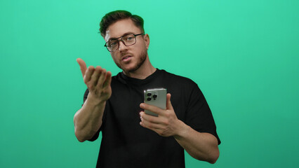 Young man with beard and glasses uses smartphone against isolated green wall background displaying various expressions with focus on technology and communication.