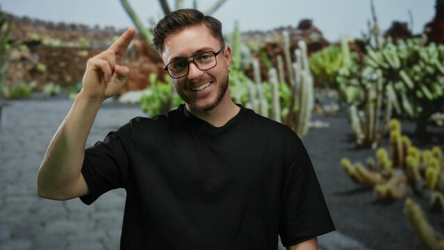 Young man with beard smiling in park setting, gesturing with fingers on forehead, wearing glasses and black shirt, surrounded by green plants and stone walls outdoor.