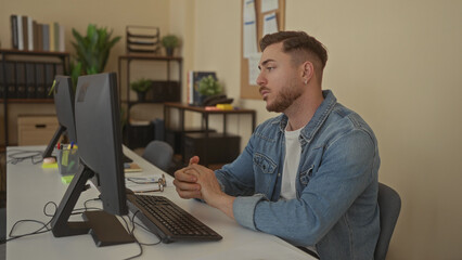 Young man with beard in office engaging in videoconference at desk with computer in professional indoor setting emphasizing modern workplace environment and communication.