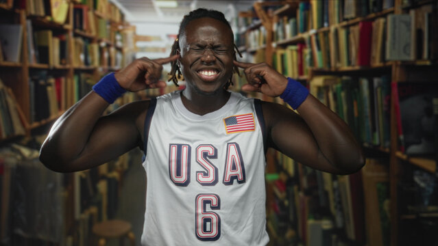 Man with fingers in ears grimacing in library wearing usa jersey and wristbands; study distraction frustration.