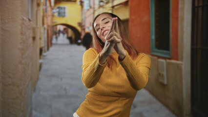 Fototapeta premium Young woman with red hair making a finger gun gesture with hands and fingers in a narrow cobbled street archway; playful confidence.