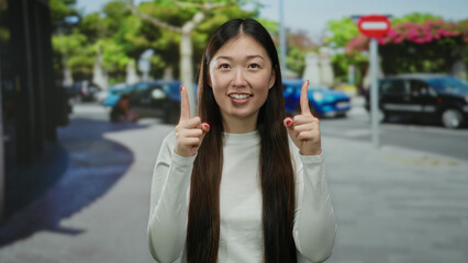 Woman smiling outdoors in urban city street setting with trees, wearing casual white attire and gesturing upwards joyfully during daytime.