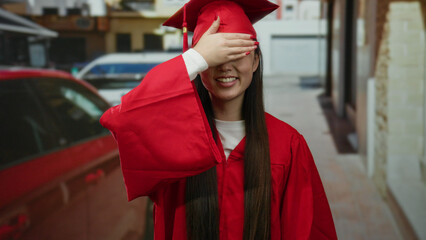 Woman in red graduation outfit playfully covering eyes on urban street celebrating academic success...