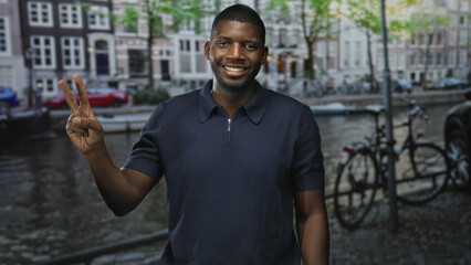 Fototapeta premium Man smiling shows two fingers with right hand beside a canal on a cobbled street, wearing a navy polo with bicycles and moored boats visible; joy travel.