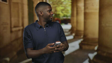 Man holding smartphone with both hands, smiling and looking to his right amid stone columns in a building; curiosity exploration.