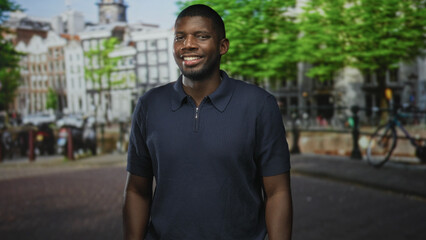 Fototapeta premium Man smiling with arms relaxed, standing on a canal street by a bicycle and railing, wearing navy polo shirt; confidence leisure.