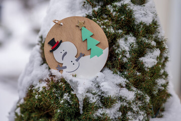 Wooden snowman ornament on evergreen tree with snow and icicle. Natural winter decoration outdoors, showing melting snow, suitable for seasonal design, winter themes, greeting cards and backgrounds.