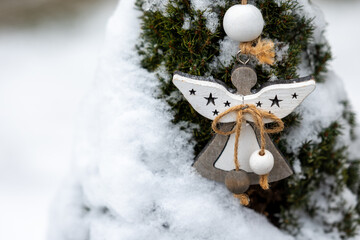 Wooden angel ornament with beads and rope hanging on a snow covered evergreen tree. Rustic winter decoration suitable for holiday backgrounds and seasonal design.