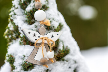 Wooden angel ornament hanging on a snow covered evergreen tree. Natural winter decoration with rope and beads, suitable for holiday backgrounds, seasonal design and greeting cards.