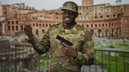 Soldier man in camouflage uniform holding a handgun at chest and pointing index finger upward in a building ruin with metal railing and stonework and grass; pride.