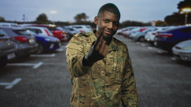Man soldier in camouflage shows middle finger with bare finger toward camera in street parking lot while smiling and leaning forward; defiance resolve.