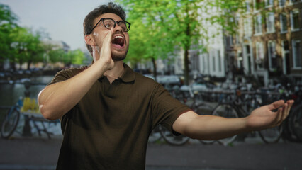 Young hispanic man cupping hand to mouth shouting toward canal and parked bicycles on amsterdam street, gesturing with open arm; urgency.