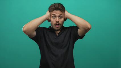 Fototapeta premium Young hispanic man with hands over ears posed in black polo in teal studio under studio lighting; surprise reaction.