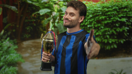 Man holding trophy and extending right hand for handshake in forest with green foliage and blue...