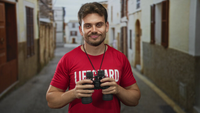 Man lifeguard in red shirt smiling and holding binoculars while standing on an old town street lined with stone buildings; duty vigilance safety.